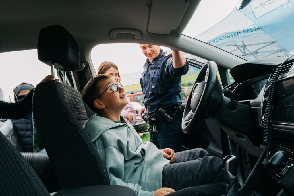 Kind sitzt am Fahrersitz des Streifenwagens, eine Polizistin schaut zu